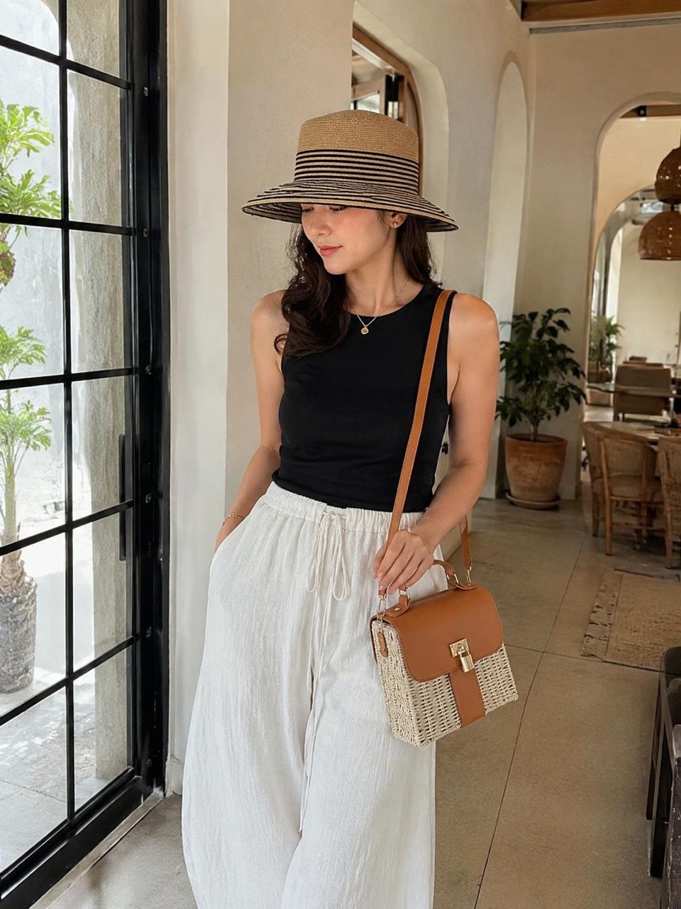 A woman by a tall window in white linen, wearing a striped woven hat with a small woven crossbody bag